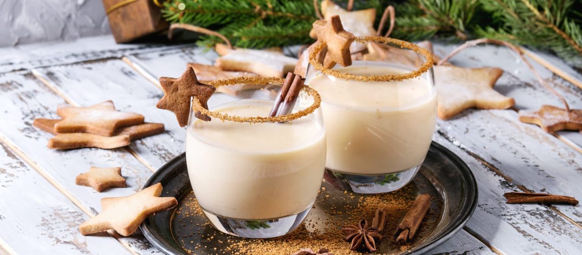 Eggnog Christmas milk cocktail with cinnamon, served in two glasses on vintage tray with shortbread star shape sugar cookies, decor toys, fir branch over white wooden plank table.