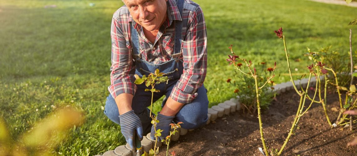 Enthusiastic senior man crouching near a flowerbed and digging in the ground while looking the camera